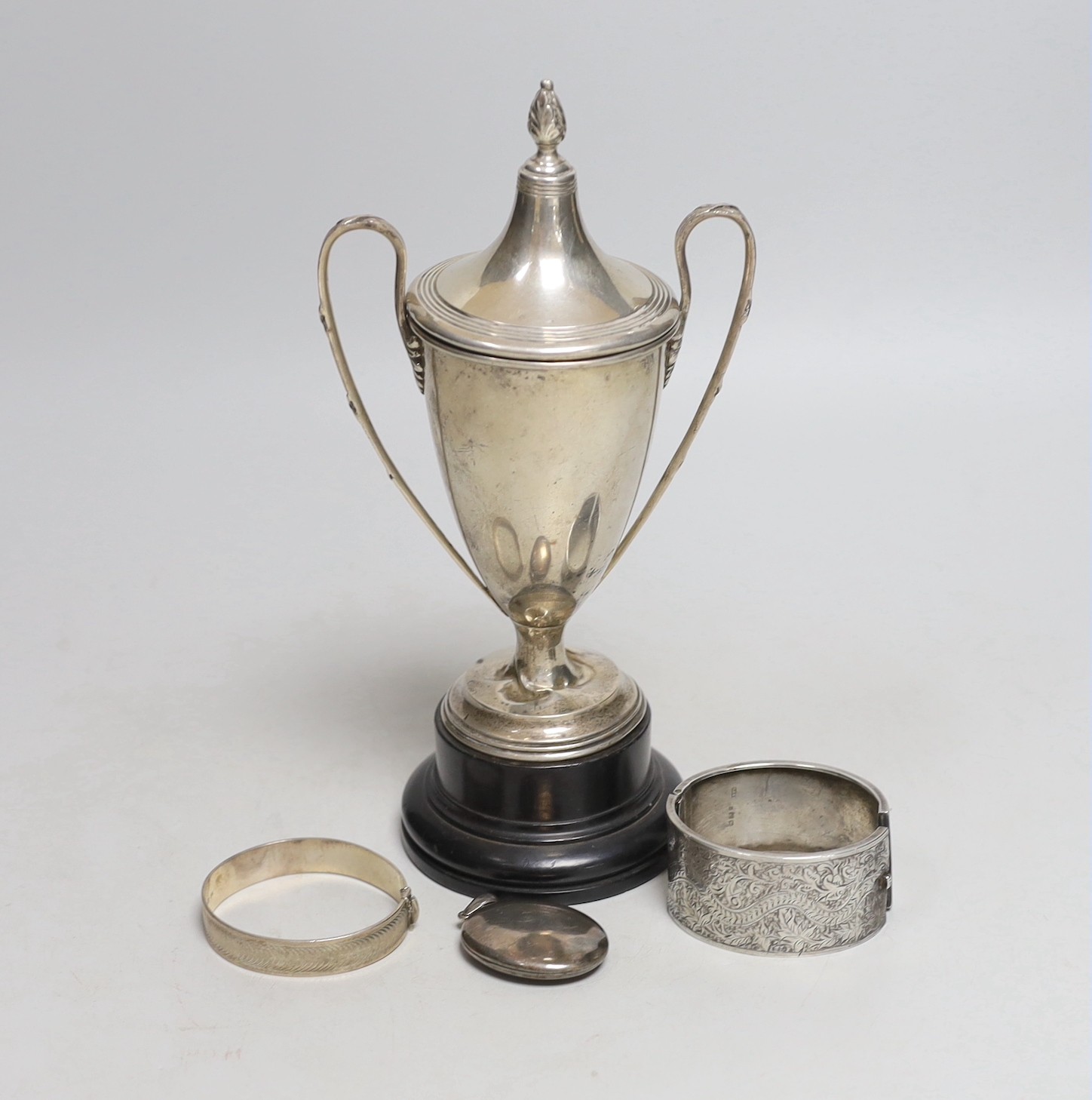 A George V silver two handled trophy cup, 19.6cm, on stand, two silver hinged bangles and a white metal locket.